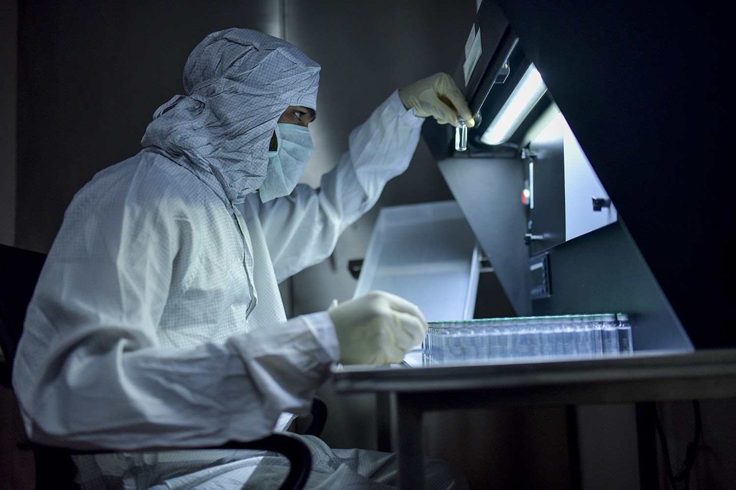 In a dark lab, a scientist in personal protective gear studies a glass vaccine vial.